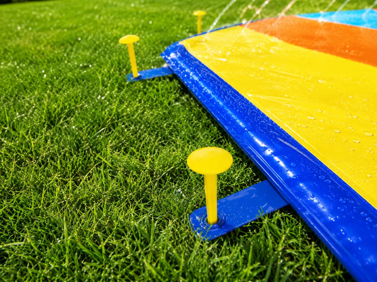 Ground pegs securing the edge of a colorful backyard slip and slide on grass while the sprinkler system sprays water